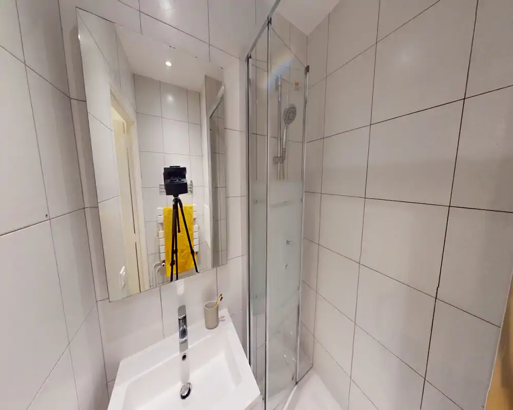 Bagno moderno compatto con piastrelle bianche, box doccia ad angolo con porta in vetro e piccolo lavabo con specchio — pulito e funzionale.