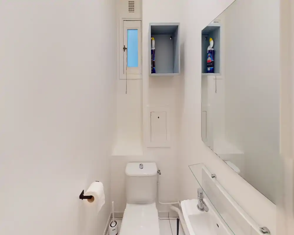 Bagno compatto e pulito con wc, piccolo lavabo e specchio a parete; finiture bianche e un nicchia a muro per lo stoccaggio rendono lo spazio essenziale e ordinato.