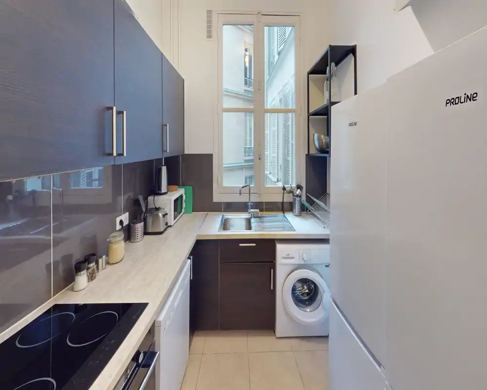 Cucina moderna e compatta con mobili scuri, piano cottura elettrico integrato, lavello sotto una finestra ampia e elettrodomestici integrati tra cui lavatrice e frigorifero.