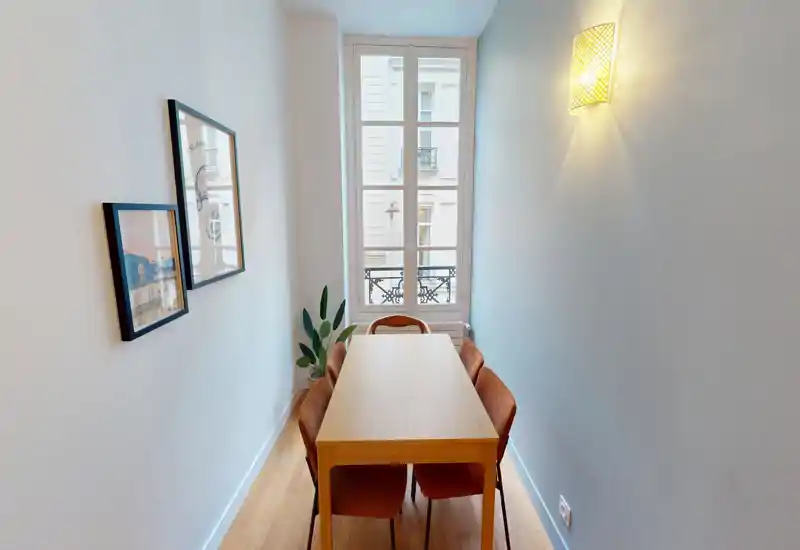 Angolo pranzo stretto e ben illuminato con tavolo in legno e quattro sedie imbottite di fronte a una finestra alta; semplici quadri a parete e una pianta completano l'allestimento minimal e accogliente.