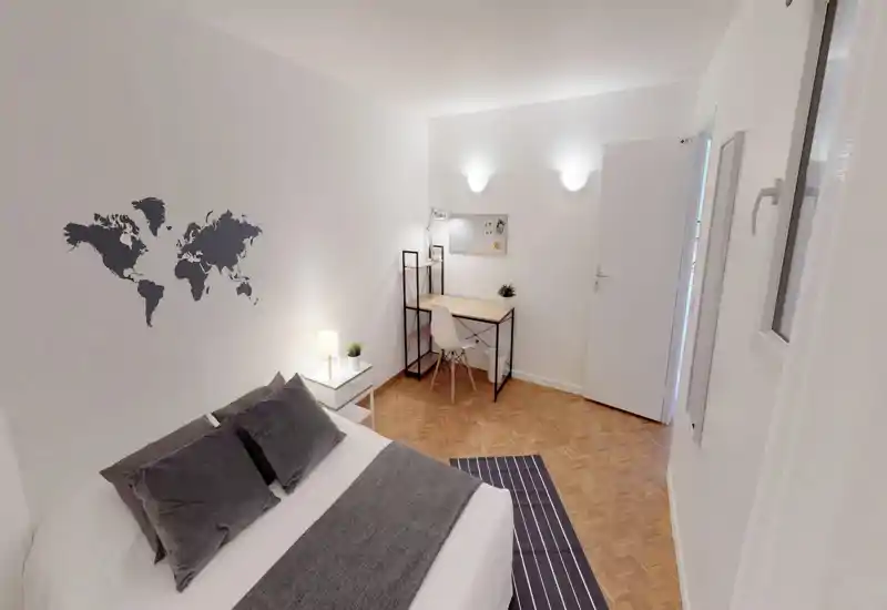Bright, modern bedroom with a double bed, bedside table with lamp and a compact desk area — neutral white walls with a world-map decal and parquet flooring create a clean, cozy atmosphere.
