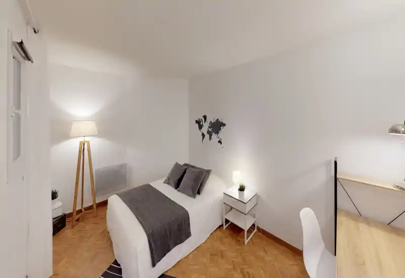 Bright, minimalist bedroom with a double bed dressed in white linens and gray accents, bedside table with lamp and a small desk area — ideal for a calm stay.