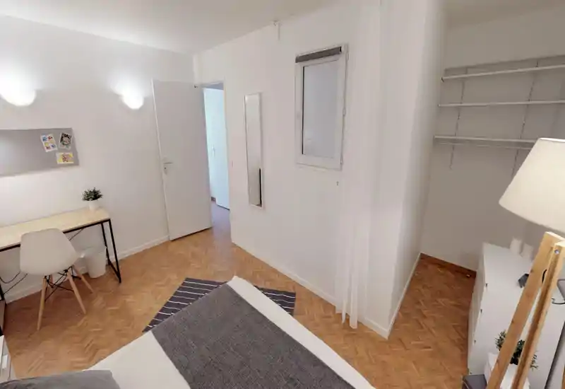 Bright, minimalist bedroom with a visible bed in the foreground, a small desk and chair, open shelving and warm parquet flooring — ideal for a single occupant or student.