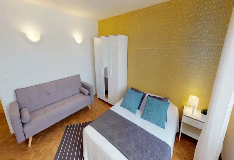 Bright, well-staged bedroom with a double bed, patterned yellow accent wall, bedside table and lamp, plus a compact sofa and wardrobe — ideal for a cozy short-term stay.