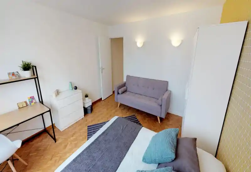 Bright, modern bedroom with a comfortable double bed in the foreground, cozy textiles and a compact sofa. The room features wood parquet floors, a study desk, dresser and pleasant neutral decor ideal for rental photos.