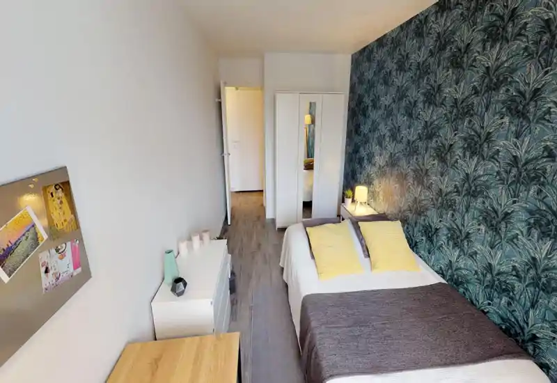 Bright, well-staged double bedroom with patterned accent wall, bedside lamp and two yellow cushions adding a cheerful touch. The bed is fully visible and the room includes wardrobe and dresser for practical storage.