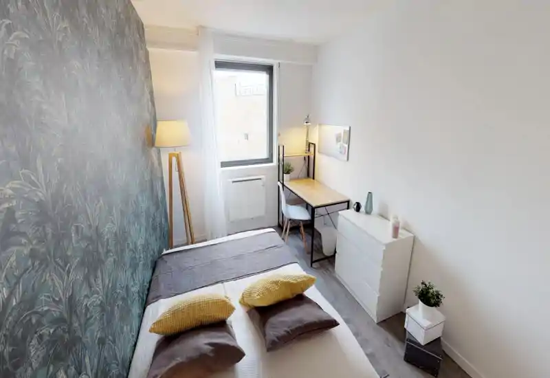 Bright, modern bedroom with a double bed, decorative pillows and a patterned accent wall. A window provides natural light and a compact desk with chair creates a functional workspace.