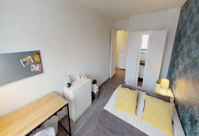 Bright, well-staged bedroom with a double bed dressed in neutral linens and yellow accent cushions, a bedside lamp, wardrobe with mirror and a small desk — ideal for a comfortable stay.