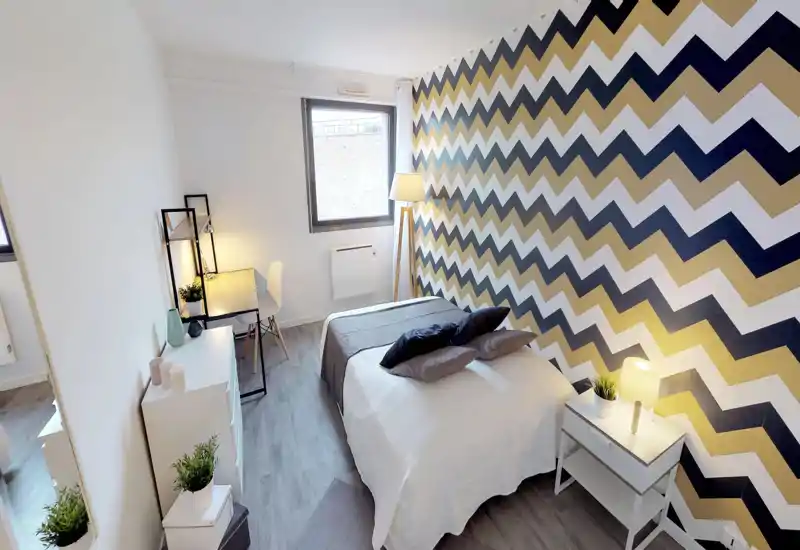Bright, modern bedroom with a double bed centered in the room, decorative zigzag accent wall, bedside table with lamp and small desk by the window — comfortable and well lit.
