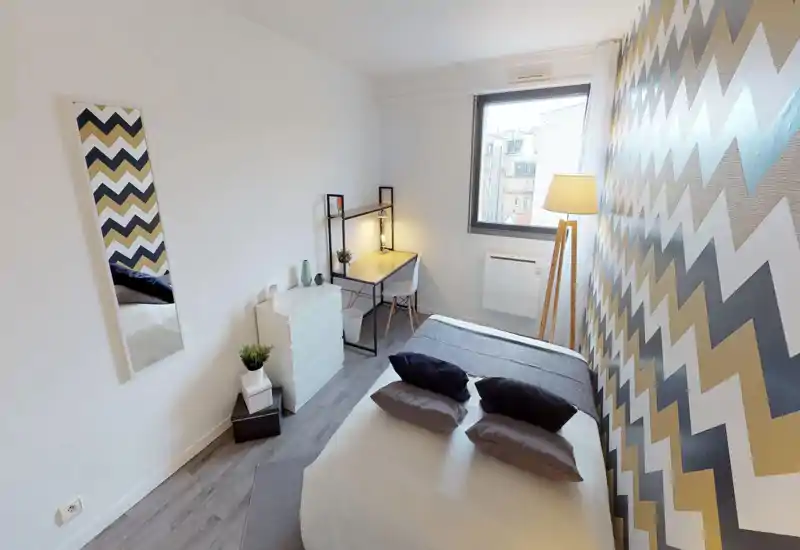 Bright, modern bedroom with a double bed, decorative pillows and a striking zigzag accent wall. The room includes a small desk by the window, a standing floor lamp and neat storage, ideal for comfortable stays or work-from-home.