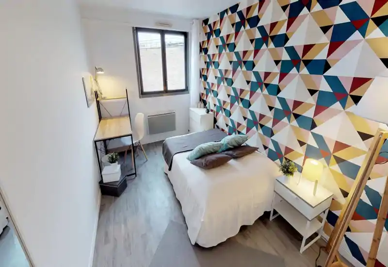 Bright, modern bedroom with a double bed, decorative pillows and a geometric accent wall; includes a bedside table with lamp and a compact desk by the window.