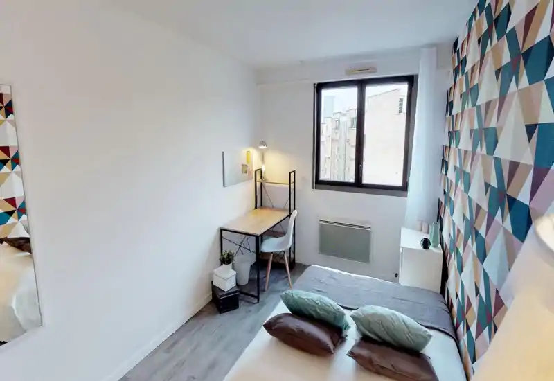 Bright, modern bedroom with a double bed in the foreground, decorative pillows and a geometric accent wall; a small desk by the window adds a functional workspace.