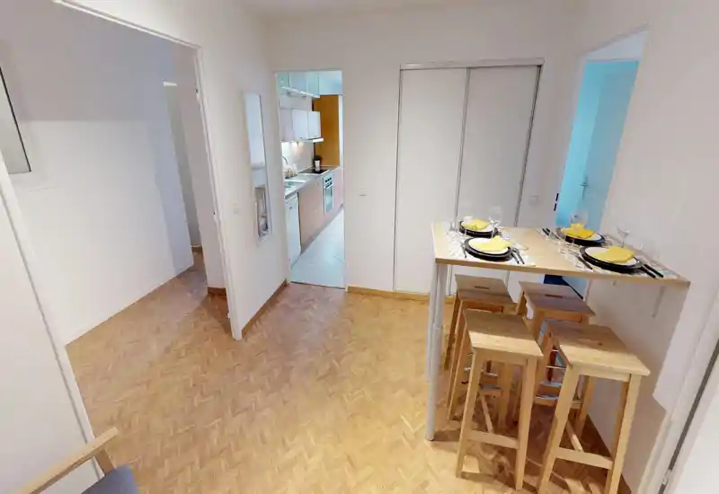 Compact, modern living/dining corner with a raised wooden breakfast bar and stools; bright, clean parquet flooring and an open doorway leading to the kitchen.