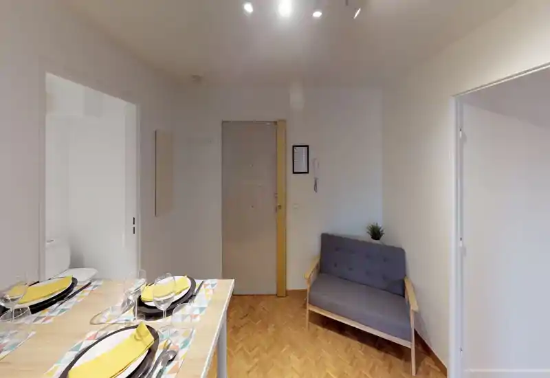 Compact, well-lit living area with a two-seater sofa, small decorative plant and a dining counter set for four — ideal for a cozy studio layout.