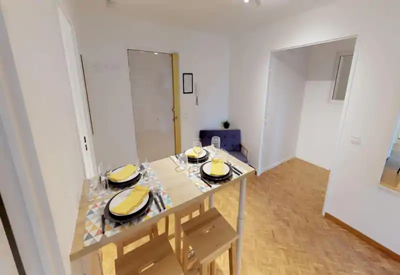 Bright, neatly staged dining area with a wooden high table set for four, colorful placemats and stools; light parquet flooring and a small seating bench create a welcoming spot for meals.