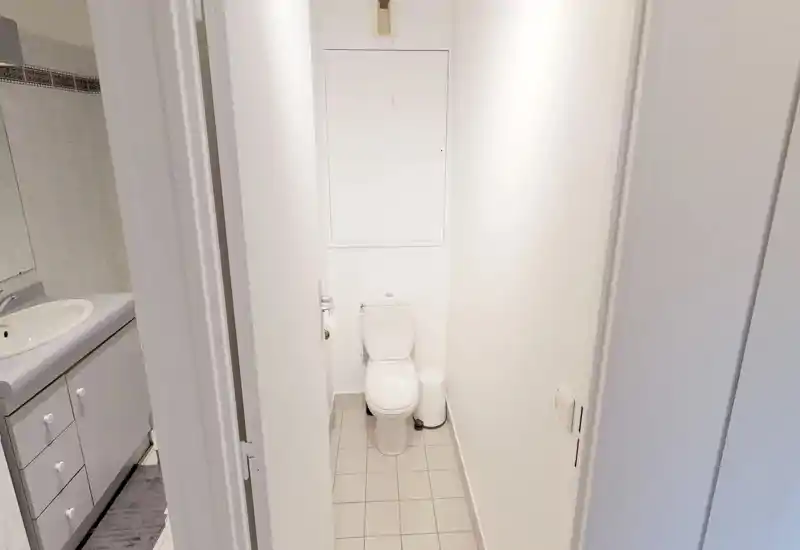 Compact bathroom with white tiles and a standard toilet; a sink and vanity are visible through the doorway. Clean, bright and simple layout suitable for a small apartment.