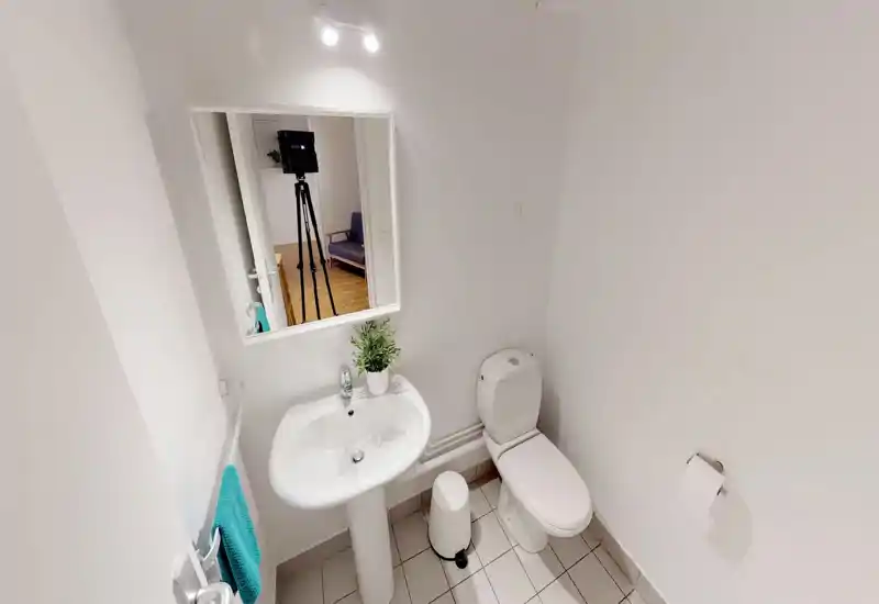 Small, bright bathroom with a pedestal sink, toilet and mirror. Clean white tiles and simple fixtures create a neat, functional space ideal for short-term stays.