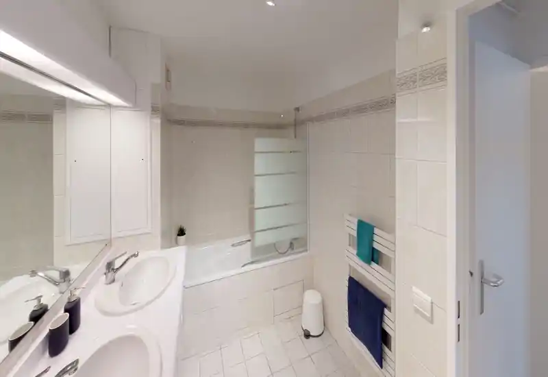 Clean, tiled bathroom with a bathtub and shower screen, double sink vanity, heated towel rail with towels, and bright even lighting.