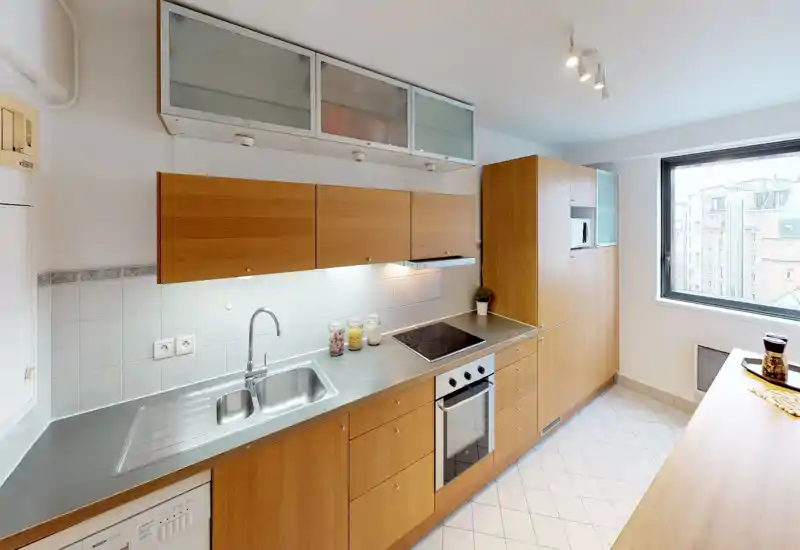 Modern, well-lit kitchen with wooden cabinets, stainless steel sink and integrated electric oven and hob. Large window provides natural light and the layout offers practical counter space for meal preparation.