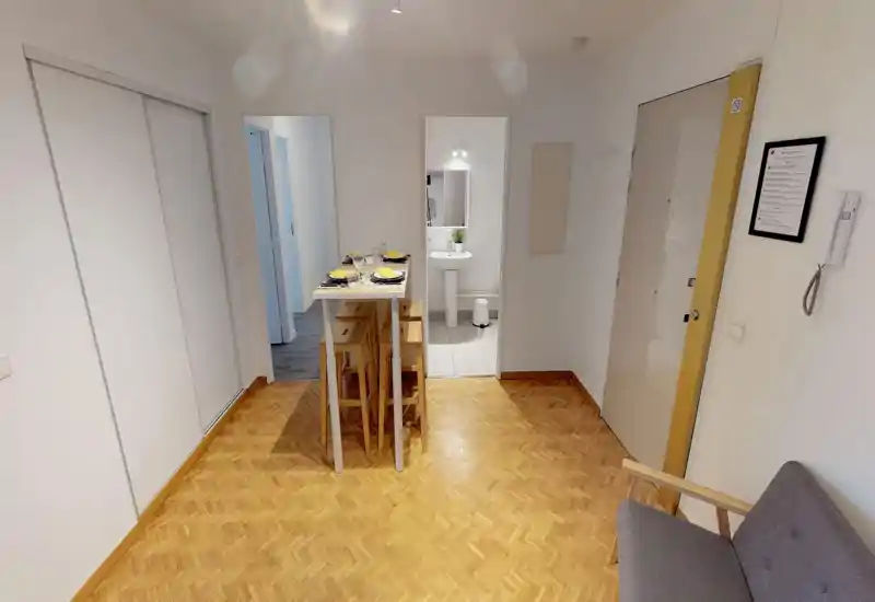 Zona living compatta e luminosa con pavimento in parquet, un piccolo divano e un tavolo alto apparecchiato per quattro persone — spazio ordinato e funzionale, ideale per soggiorni brevi.
