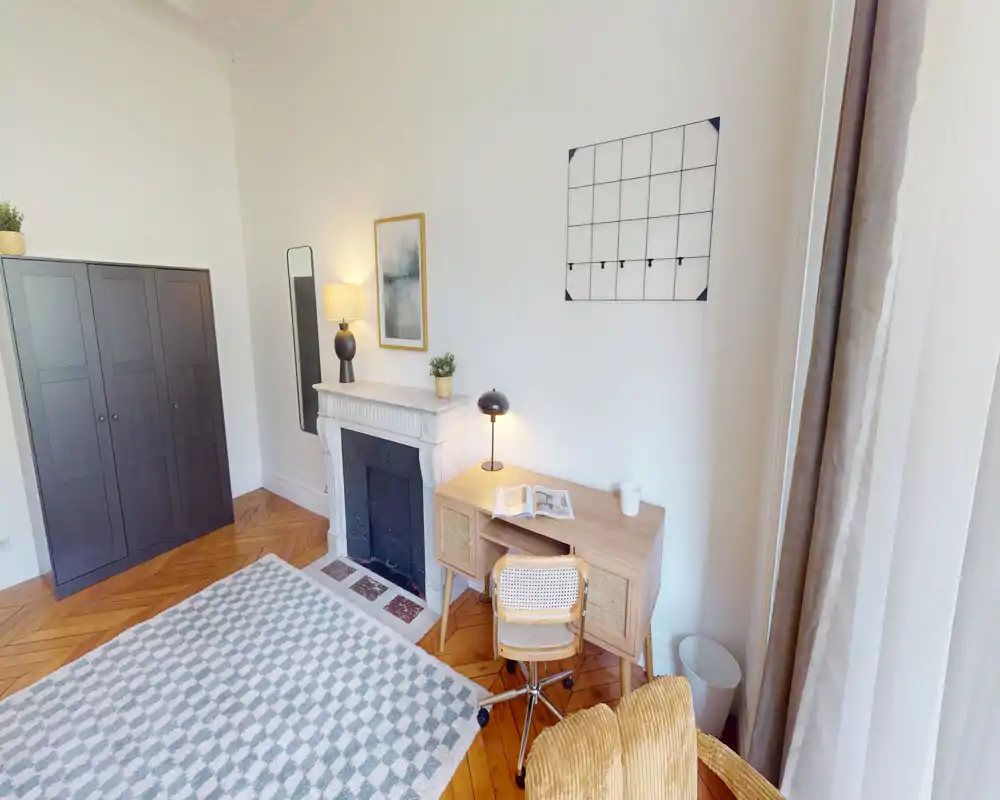 Coin de chambre lumineux et élégant avec bureau, cheminée décorative et armoire ; la lumière naturelle et le parquet en chevrons apportent une atmosphère chaleureuse.