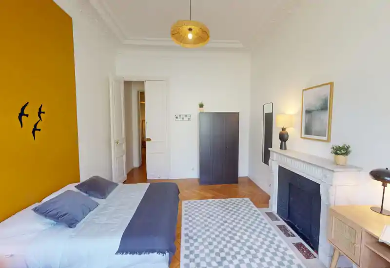 Chambre lumineuse et élégante avec lit double contre un mur moutarde, parquet, cheminée décorative et tapis à motifs — idéale pour un séjour cosy en ville.