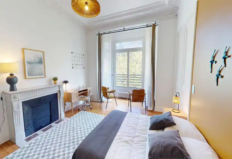 Chambre lumineuse et élégante avec lit double, coussins décoratifs, cheminée, bureau et grande fenêtre apportant une belle lumière naturelle.