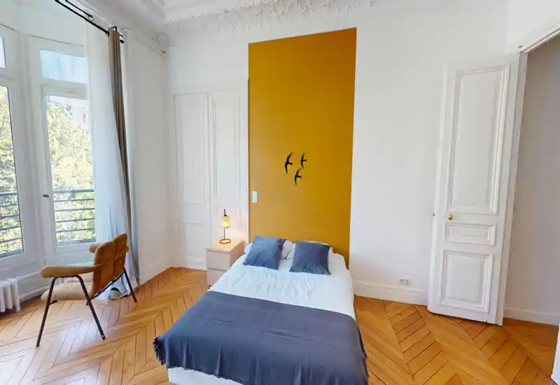 Chambre lumineuse et élégante avec un lit double, un mur d'accent moutarde et de grandes fenêtres apportant une belle lumière naturelle. Sol en parquet, lampe de chevet et fauteuil confortable près de la fenêtre.