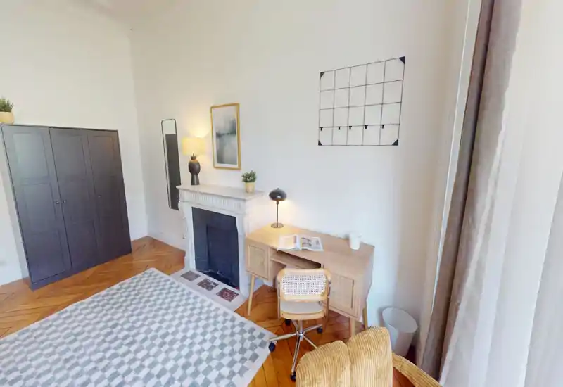 Coin de chambre lumineux et élégant avec bureau, cheminée décorative et armoire ; la lumière naturelle et le parquet en chevrons apportent une atmosphère chaleureuse.
