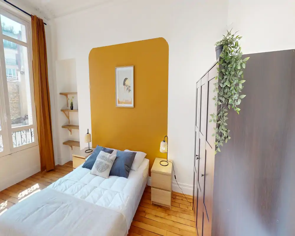 Camera da letto luminosa e curata con letto matrimoniale davanti a una parete d'accento ocra, comodini con lampade, pavimento in legno e una grande finestra che offre molta luce naturale.