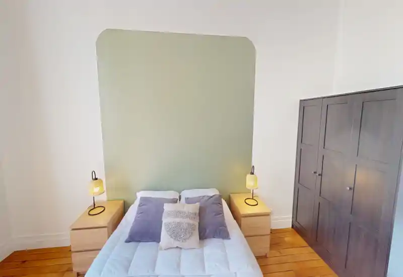 Chambre lumineuse et bien aménagée avec un lit double centré, coussins assortis et tables de chevet avec lampes ; parquet chaleureux et une tête de lit vert pâle pour une ambiance calme et accueillante.