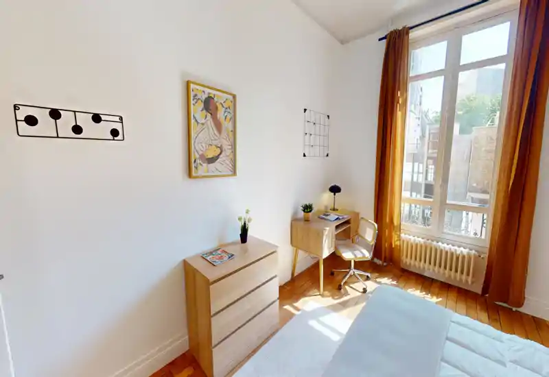 Chambre lumineuse et chaleureuse avec parquet, lit bien fait, petit bureau près d'une grande fenêtre et rideaux moutarde qui laissent entrer une belle lumière naturelle.