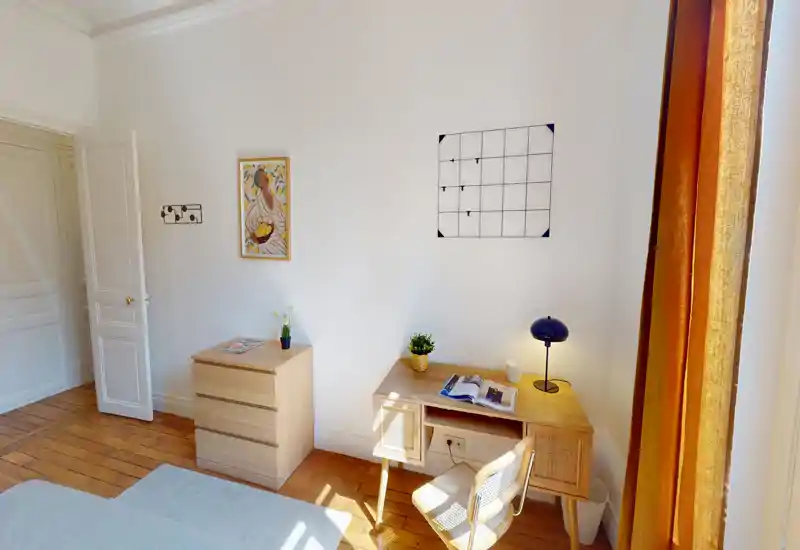 Coin de chambre lumineux et sobrement meublé avec un petit bureau en bois, une chaise et une commode près de rideaux chauds ; une partie du lit est visible au premier plan.