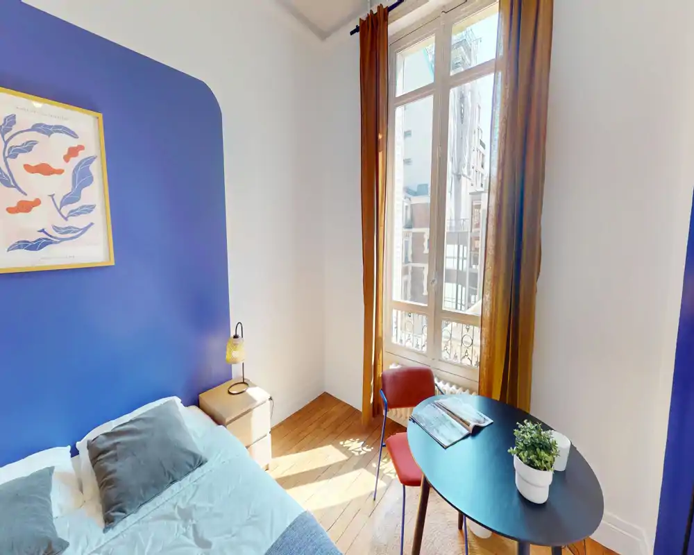 Chambre lumineuse et chaleureuse avec un mur bleu, lit double avec oreillers, table de chevet et lampe, et une petite table près d'une grande fenêtre ensoleillée.
