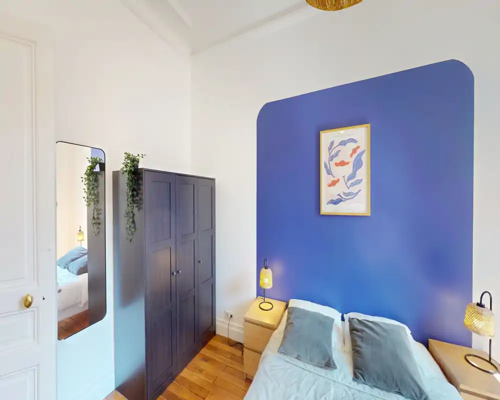 Chambre confortable et soignée avec un lit double devant une tête de lit peinte en bleu, deux tables de chevet avec lampes, une grande armoire sombre et une glace pleine longueur. Lumière naturelle et parquet chaleureux donnent une atmosphère accueillante.