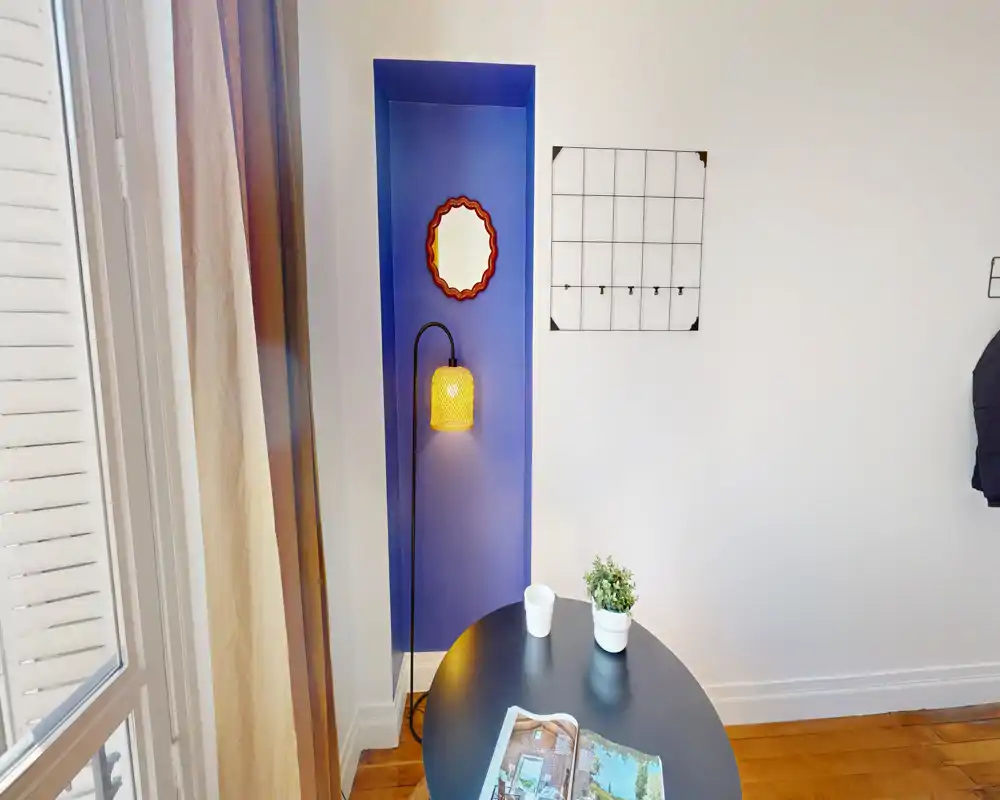 Coin d'un séjour avec une petite table ronde, une lampe sur pied décorative dans une niche bleue, un miroir et une plante en pot — lumineux, minimal et contemporain.