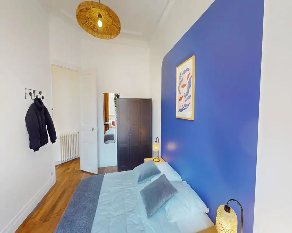 Chambre lumineuse et chaleureuse avec un mur bleu, lit double habillé de linge clair, lampes de chevet et armoire. Parquet et hauteurs sous plafond offrent une atmosphère aérée et soignée.
