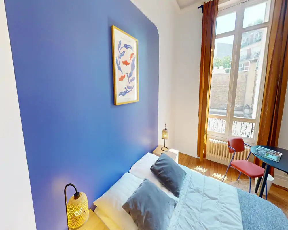 Chambre lumineuse et stylée avec un mur bleu, lit double en linge blanc et coussins gris, lampes chaleureuses et grande fenêtre apportant une belle lumière naturelle.