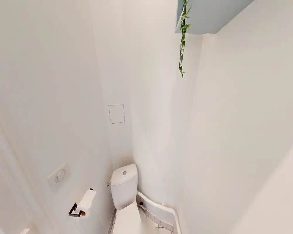 Coin de salle de bain compact avec vue rapprochée des toilettes, sol et murs carrelés blancs et un porte-papier simple ; lumineux, minimal et fonctionnel.