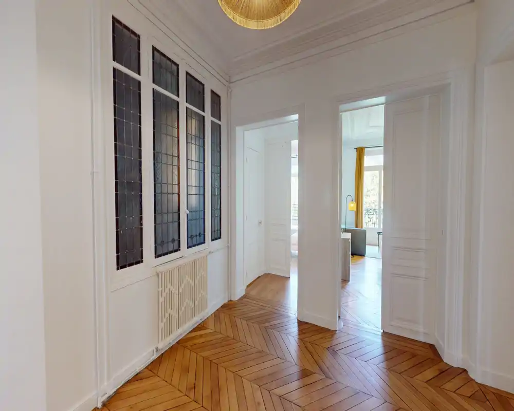 Entrée/couloir lumineux et élégant avec parquet en chevrons, murs blancs et panneaux de verre décoratifs ; dessert des pièces adjacentes baignées de lumière.