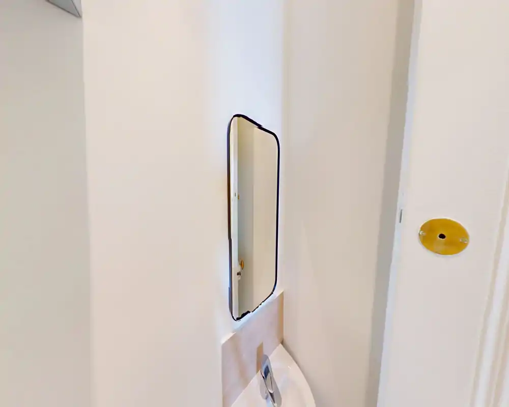 Détail de salle de bain : petit miroir mural au-dessus d’un lavabo compact, murs blancs et robinetterie simple.