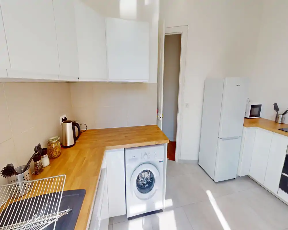 Cuisine moderne et lumineuse avec placards blancs, plans de travail en bois, réfrigérateur, micro-ondes et lave-linge sous le plan; agencement propre et fonctionnel éclairé naturellement.