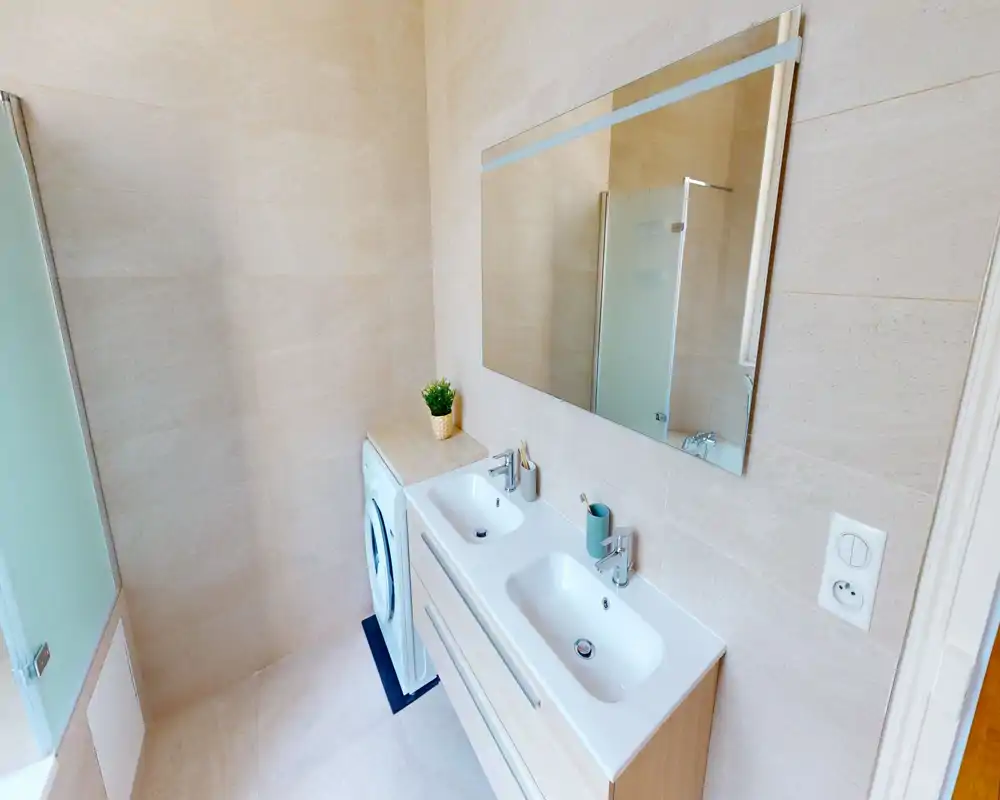 Salle de bains moderne et lumineuse avec double vasque, grand miroir et porte de douche en verre; carreaux clairs et machine à laver à côté du meuble.
