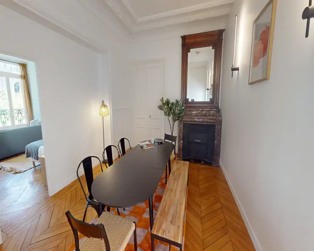 Salle à manger spacieuse avec une grande table noire, sièges variés (chaises et banc), parquet et cheminée décorative — espace lumineux et stylé, idéal pour les repas à plusieurs.