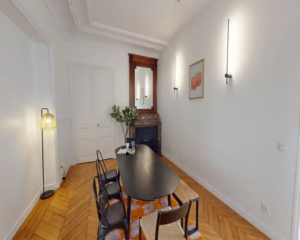 Salle à manger lumineuse et élégante avec une grande table noire, chaises assorties, tapis décoratif et une cheminée classique avec miroir — idéale pour dîners et réunions.