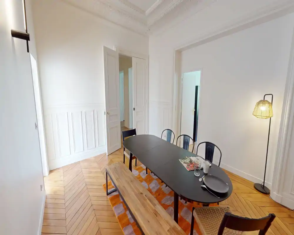 Salle à manger lumineuse et élégante avec une grande table noire, une banquette, des chaises en métal et une lampe sur pied ; le parquet point de Hongrie et le tapis décoratif apportent une atmosphère chaleureuse pour les repas.