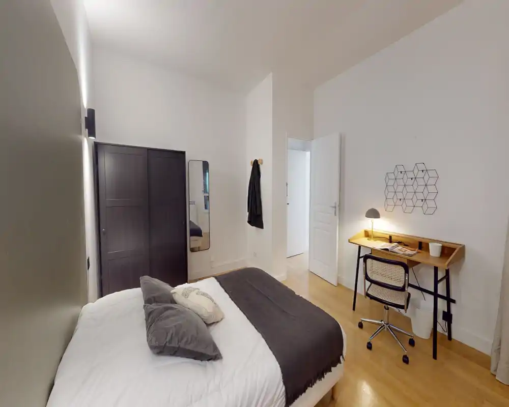 Chambre lumineuse et soignée avec un lit double en linge blanc et accents gris, une armoire foncée et un petit bureau avec lampe — idéale pour travailler et se détendre.