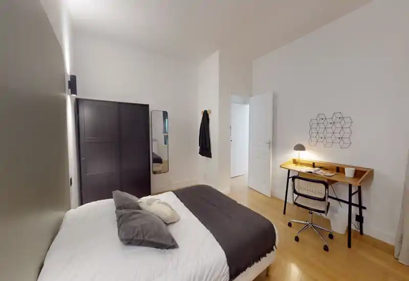 Chambre lumineuse et soignée avec un lit double en linge blanc et accents gris, une armoire foncée et un petit bureau avec lampe — idéale pour travailler et se détendre.