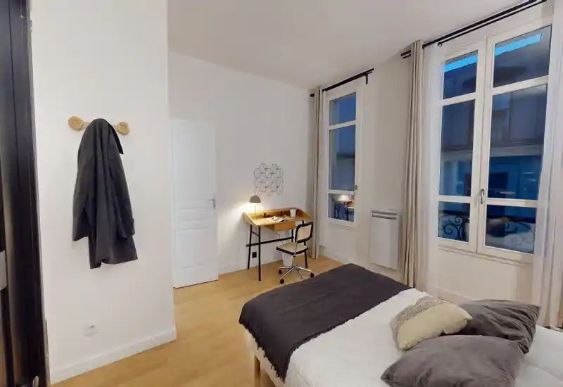Chambre calme et soignée avec lit double, coussins confortables et petit bureau en bois près de la fenêtre. Grandes fenêtres apportant une belle luminosité et une décoration neutre qui crée une atmosphère apaisante, parfaite pour une annonce.