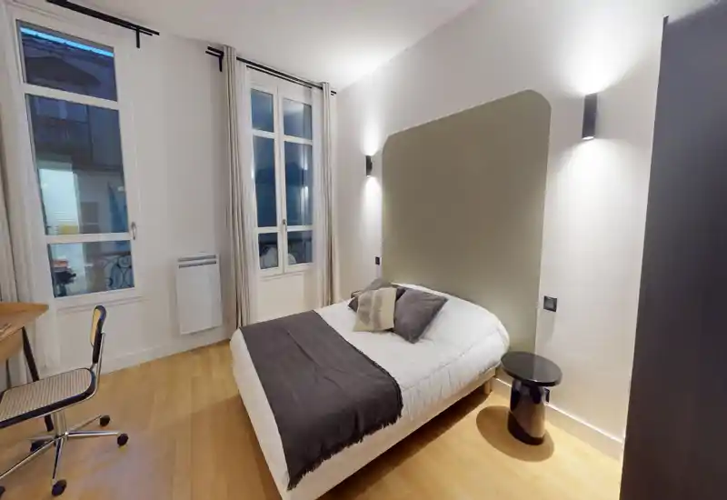 Chambre moderne et lumineuse avec un lit double centré, table de chevet et appliques murales; parquet clair et grandes fenêtres apportant luminosité et confort.